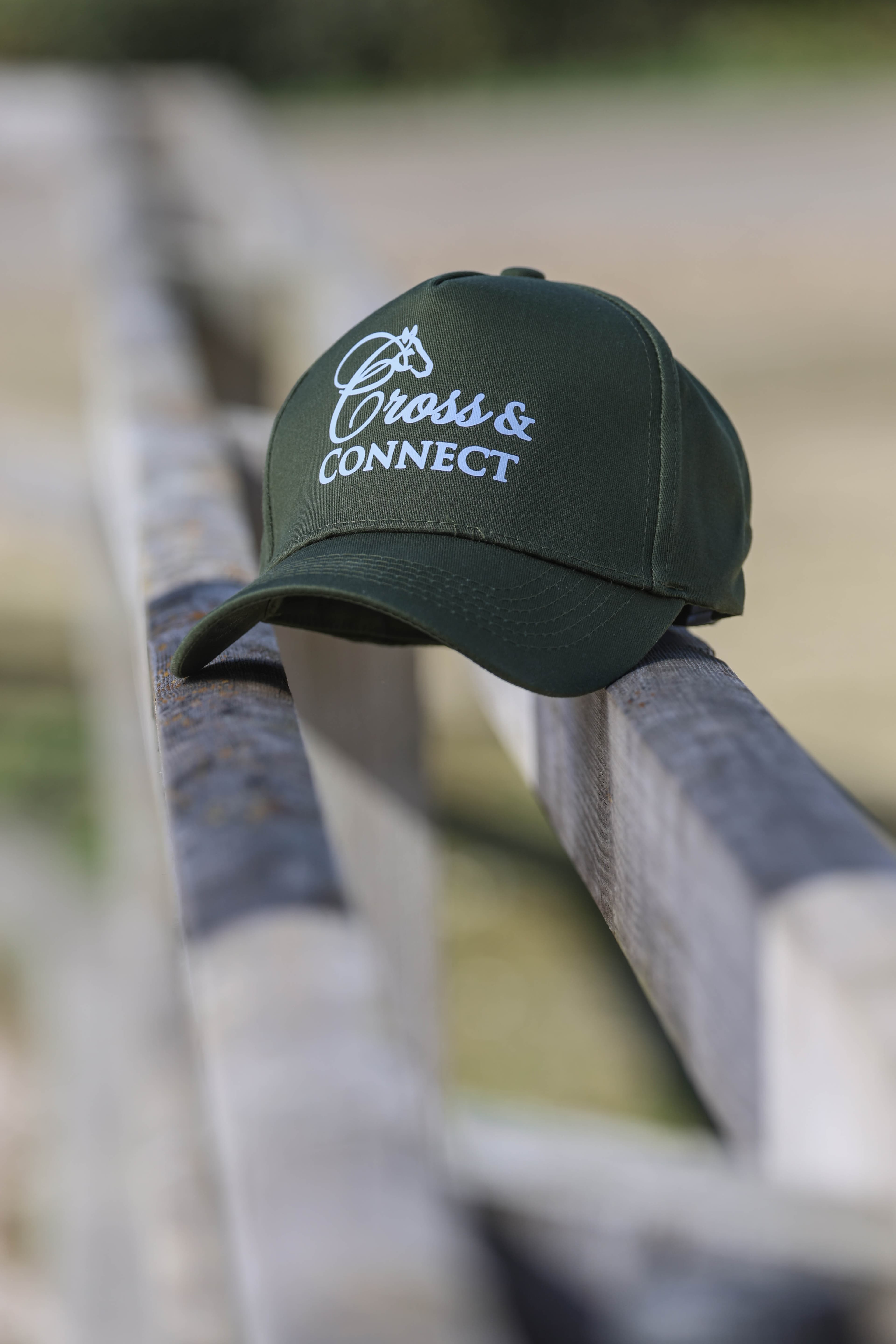 A dark green Cross & Connect cap resting on a wooden fence beside the course.